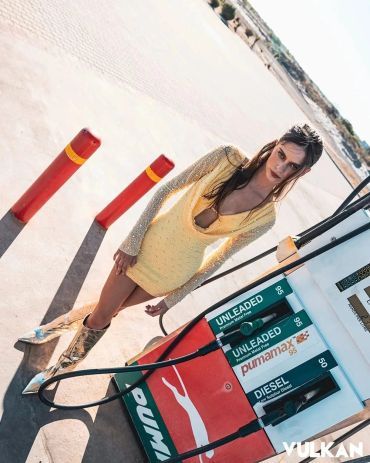 Woman in yellow dress stands by gas pump at sunny station.