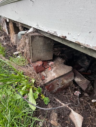 Close-up of a house foundation with bricks and wooden support, surrounded by grass and soil.