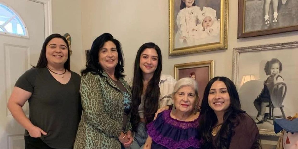 Five women of different ages smiling indoors with vintage family portraits in the background.