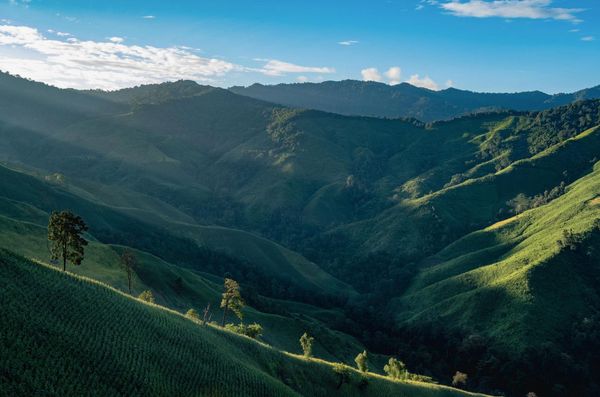Sunlight casts shadows over rolling green hills and distant mountains under a blue sky.