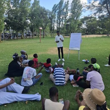 A man teaches a group outdoors with a flip chart on a grassy field.