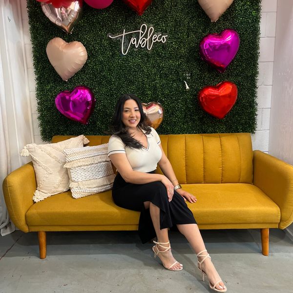 Woman sitting on mustard yellow couch with heart-shaped balloons and greenery backdrop.