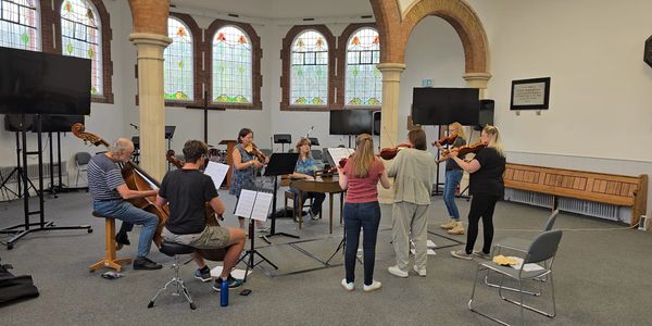 A group of musicians rehearsing in a spacious room with stained glass windows.