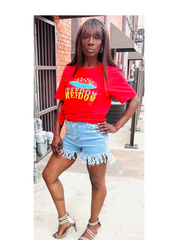 Woman in a red 'Freedom' shirt and denim shorts posing confidently outdoors.