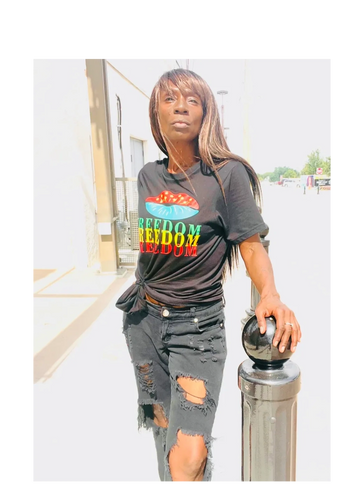 Woman in a black "Freedom" t-shirt and ripped jeans leaning on a post outdoors.
