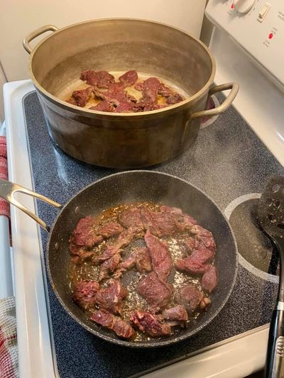 Caribou meat cooks in a pot & pan on a stovetop, steam is visible and a mixing spoon sits nearby. 