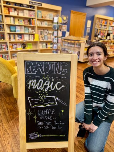 Young woman smiles beside a bookstore sign promoting reading magic and hours.