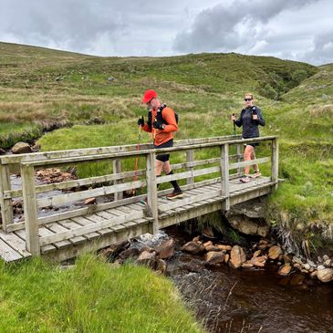 Two hikers crossing a wooden bridge over a small stream.
