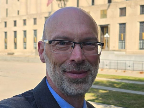 Smiling man with glasses and beard outdoors near a building.