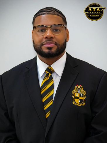 Man in a black suit with a striped tie and crest on the blazer.
