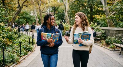Two female students walking and chatting on a campus path holding books.