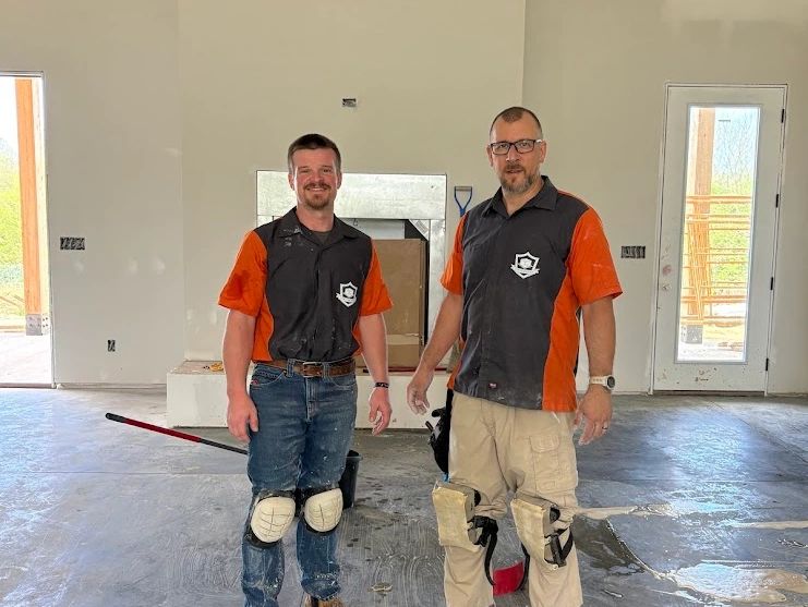 Two workers in black and orange shirts standing indoors on a concrete floor.