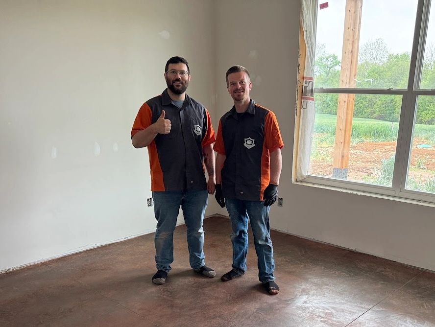 Two men in matching black and orange shirts stand in an empty room with a large window.