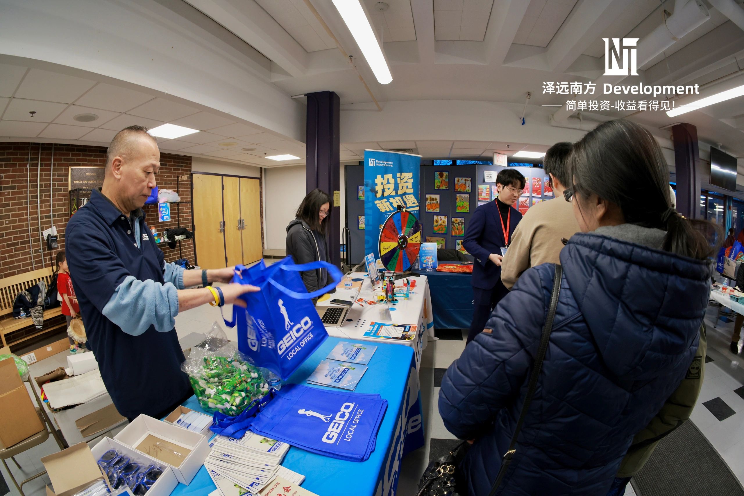 People interacting at a promotional booth with GEICO bags and a spin wheel.