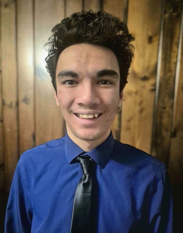 Young man smiling, dressed in blue shirt and black tie in front of wooden wall.