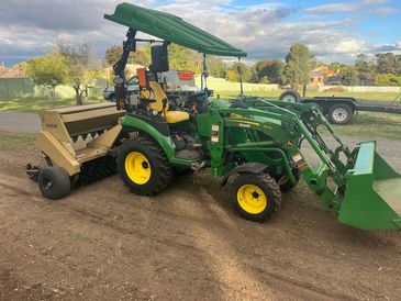Green tractor with front loader and attached seeder on dirt ground.