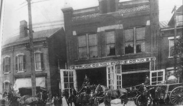 Old black and white photo of horse-drawn fire trucks outside a firehouse.