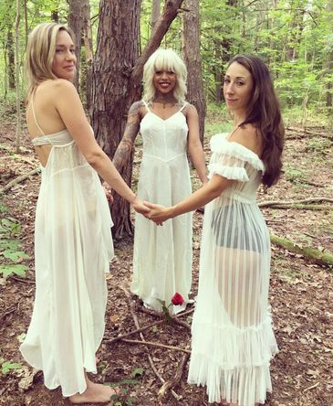 Three women in white dresses holding hands in a forest.