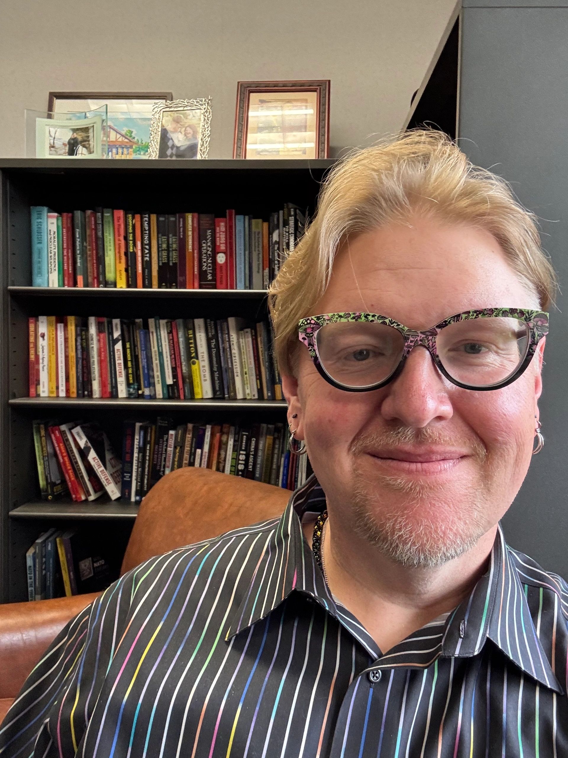 Smiling person wearing patterned glasses and a striped shirt in front of a bookshelf.