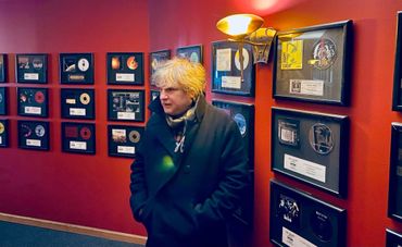 Person with blond hair stands in a room with numerous framed awards on red walls.