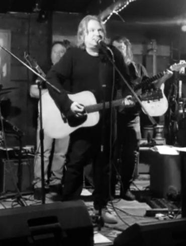 Musician performing with guitar on stage in a dimly lit venue.
