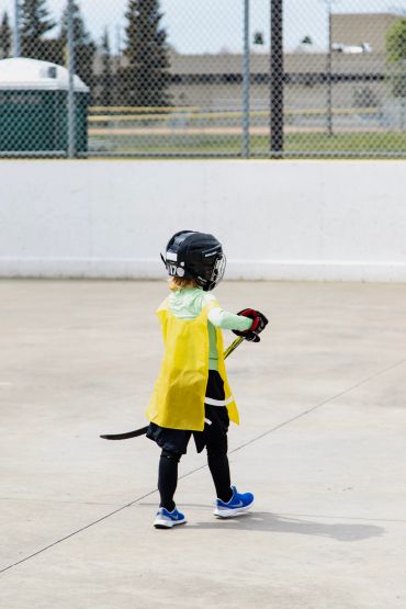 Child in hockey gear with yellow cape on outdoor rink.