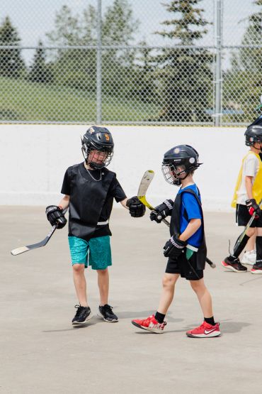 Two young boys playing street hockey outdoors, wearing helmets and sports gear.