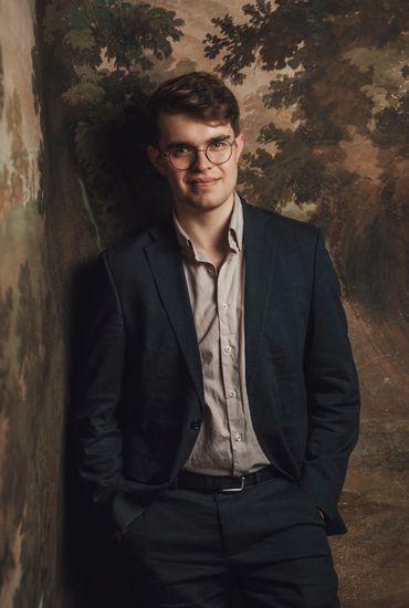 Young man in glasses wearing a suit jacket stands against a painted wall.