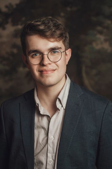 Young man with glasses in a blazer smiling gently.