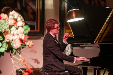Man in a suit playing grand piano under a lamp.
