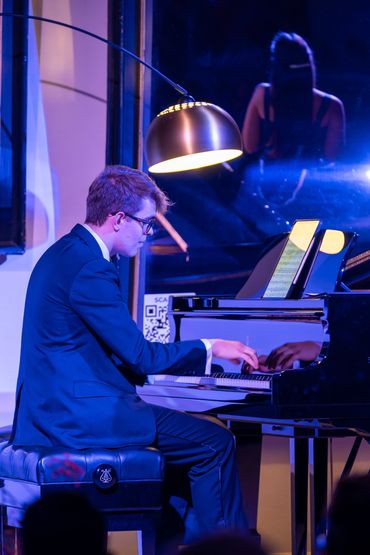 Man in a suit playing piano under a stylish lamp in a dimly lit room.