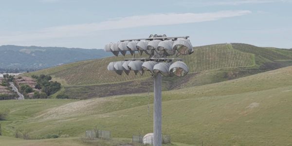 A tall pole with multiple curved floodlights in a hilly landscape.