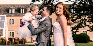 Sepia toned colour wedding photograph of a bride and groom with a young bridesmaid jumping into the 