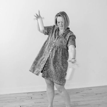 Woman joyfully dancing barefoot in a loose dress on wooden floor.