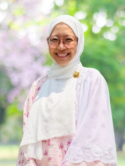 Smiling woman in a white hijab and glasses outdoors with a floral background.
