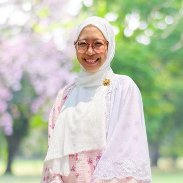 A smiling woman wearing a white hijab and glasses outdoors.