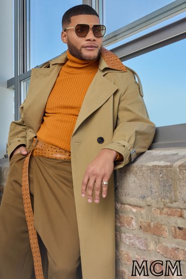Stylish man wearing a tan trench coat and sunglasses leaning on a brick wall.