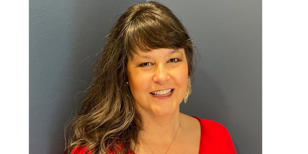 Smiling woman with long hair in a red top against a gray cornered wall.