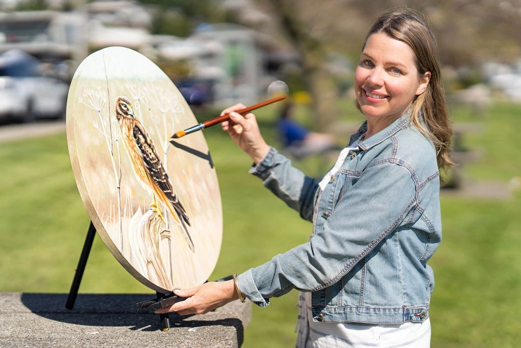 The Illustrator stands in the sun smiling as she paints a picture of a hawk. 