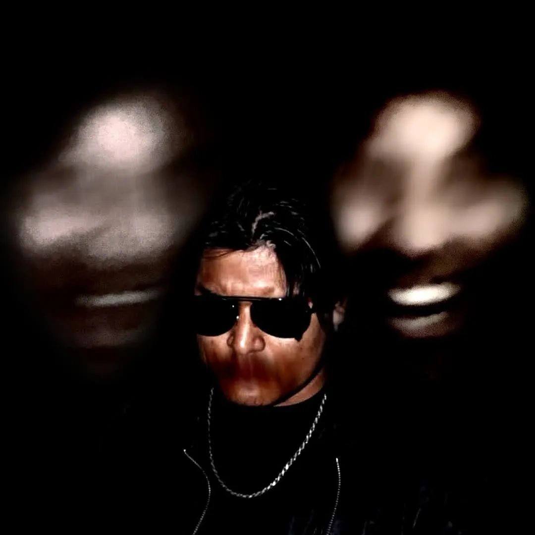 Man in sunglasses with two blurred faces in the background, dark and mysterious.