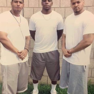 Three men in white shirts and gray shorts posing outdoors against a brick wall.