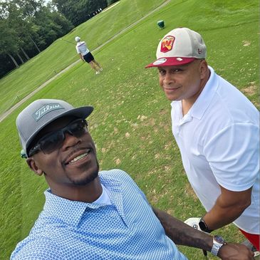 Two men smiling on a golf course with another golfer preparing to swing in the background.