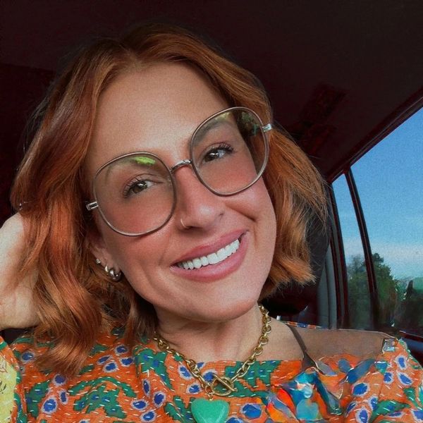 Smiling woman with red hair and large glasses wearing a colorful patterned top and green heart necklace.
