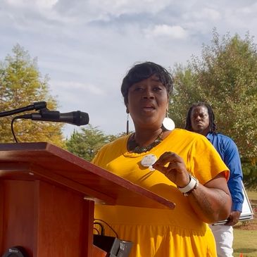Woman in yellow dress speaking at a podium outdoors.