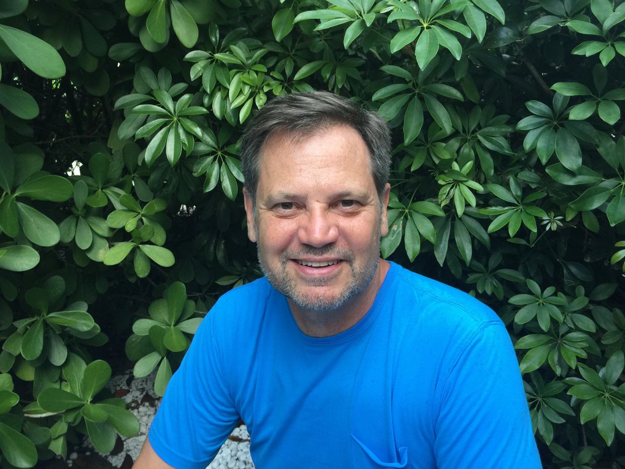 Smiling man in a blue shirt outdoors surrounded by green foliage.