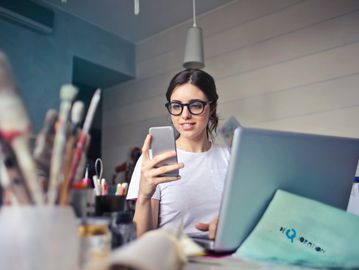 Young woman using smartphone and laptop in a creative workspace.