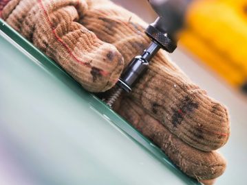 A gloved hand uses a power tool to drill into a green surface.