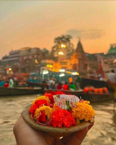 Hand holding a floral offering basket near a river at sunset.