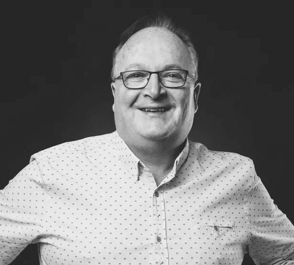 Smiling middle-aged man in glasses and patterned shirt against a dark background.