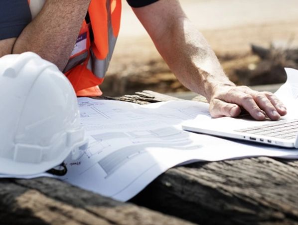 Construction worker reviewing plans with laptop and helmet on site.